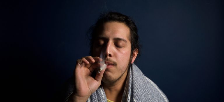 A millenial young man wearing a scarf on his shoulder deeply immersed in his smoking of cannabis.