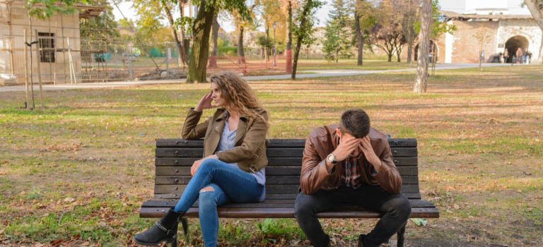 A male and female couple sitting on a bench. Both are sitting apart with the womanfacing opposite side and looking unhappy. the man is facing down and stressed. they are have a huge stressful relationship, lack of communicationand wishing they can find a solution on how to recover their relationship. this is really stressing them out.