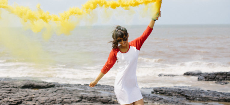 a young confident and happy lady at a beach. she is wear a gwhite gown wih red coloured sleeve. she is happy and holding a bottle filled with yellow paint spraying it into the air as she jumps.